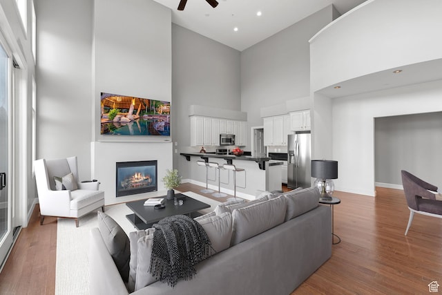 living room featuring a glass covered fireplace, a towering ceiling, baseboards, a ceiling fan, and wood finished floors
