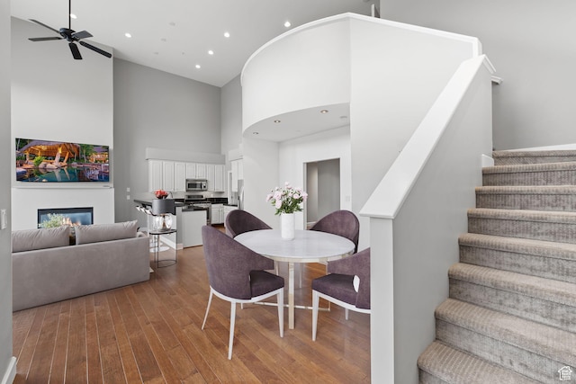 dining area featuring ceiling fan, a glass covered fireplace, hardwood / wood-style floors, a high ceiling, and stairs