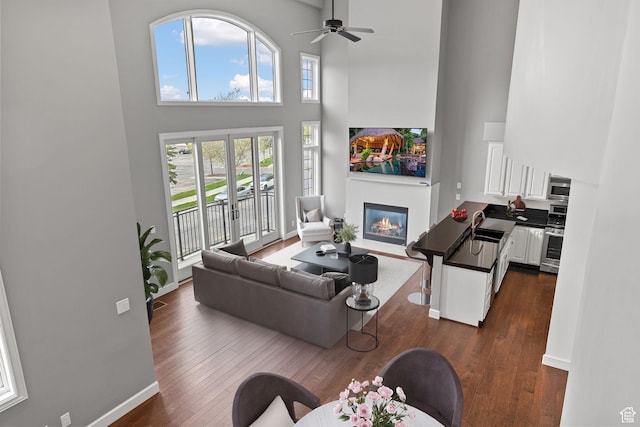 living room with baseboards, a high ceiling, ceiling fan, dark wood finished floors, and a glass covered fireplace