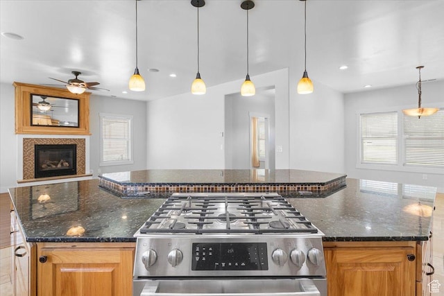 kitchen featuring stainless steel gas stove, dark stone counters, a ceiling fan, a glass covered fireplace, and open floor plan