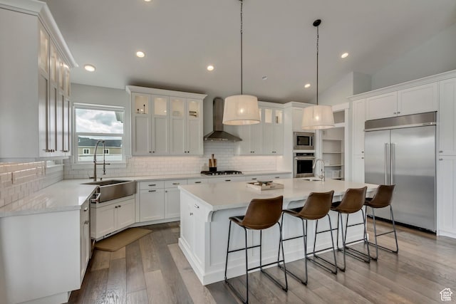 kitchen with built in appliances, an island with sink, dark wood-style floors, wall chimney exhaust hood, and a sink
