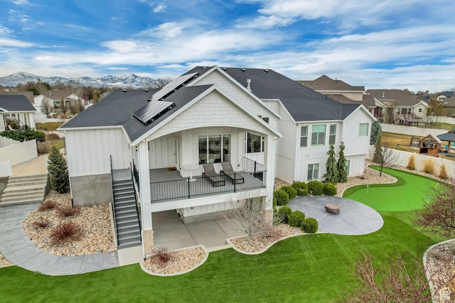 rear view of property featuring roof with shingles, board and batten siding, stairs, a patio area, and roof mounted solar panels