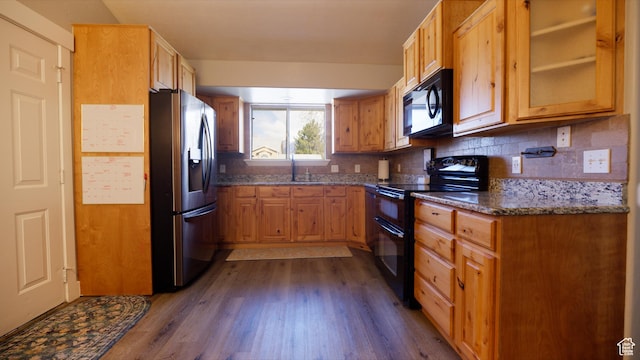 kitchen with dark stone countertops, dark wood-style flooring, a sink, black appliances, and decorative backsplash