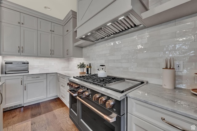 kitchen with custom exhaust hood, tasteful backsplash, wood finished floors, range with two ovens, and light stone counters