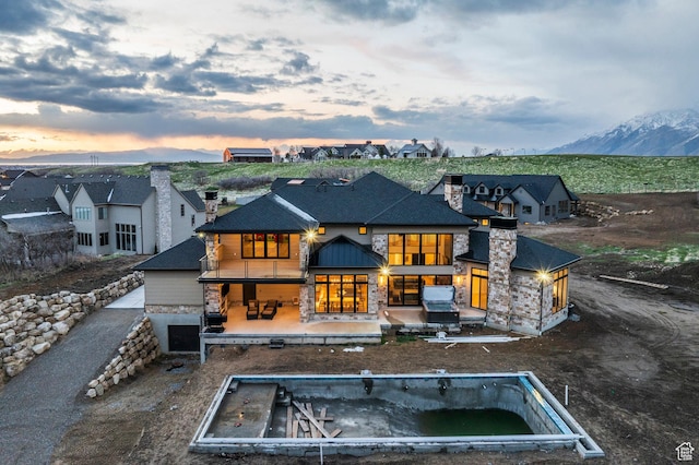 back of property at dusk with a mountain view, a hot tub, a patio area, stone siding, and a chimney