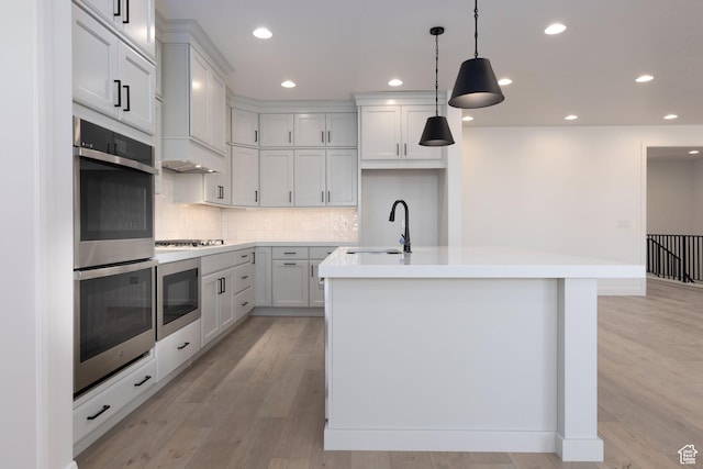 kitchen with light wood-style floors, an island with sink, gas stovetop, a sink, and stainless steel double oven