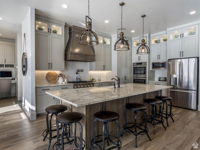 kitchen featuring a kitchen island with sink, appliances with stainless steel finishes, wood finished floors, a sink, and premium range hood