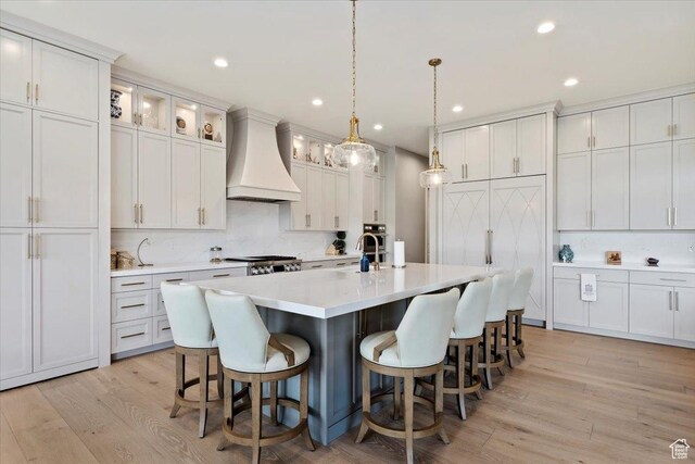 kitchen with a kitchen island with sink, custom exhaust hood, a kitchen bar, light wood-type flooring, and light countertops