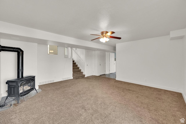 unfurnished living room featuring ceiling fan, a wood stove, visible vents, carpet flooring, and stairs