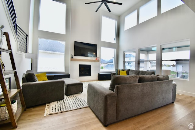 living room featuring light wood-style floors, ceiling fan, a glass covered fireplace, a high ceiling, and baseboards
