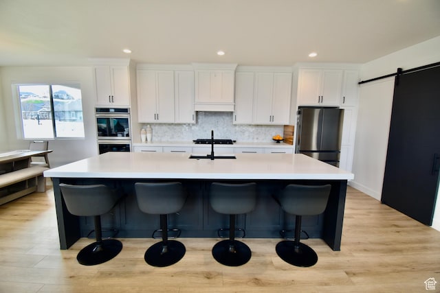 kitchen with white cabinets, a barn door, appliances with stainless steel finishes, an island with sink, and backsplash