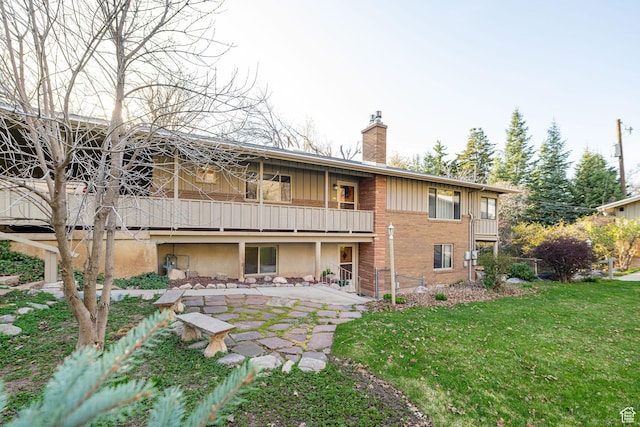 rear view of house featuring a balcony, a chimney, a patio area, brick siding, and a yard
