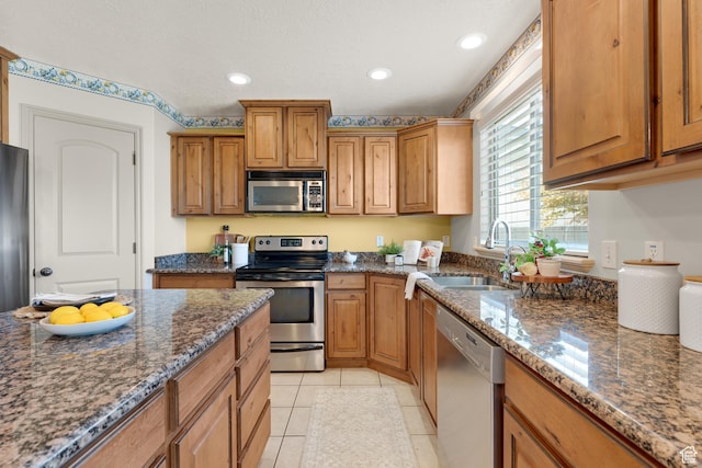 kitchen with dark stone countertops, light tile patterned flooring, brown cabinetry, stainless steel appliances, and a sink