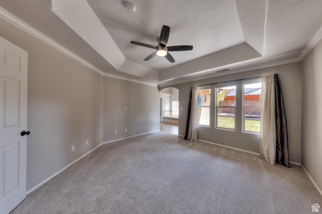carpeted spare room featuring a tray ceiling, arched walkways, ornamental molding, a ceiling fan, and baseboards