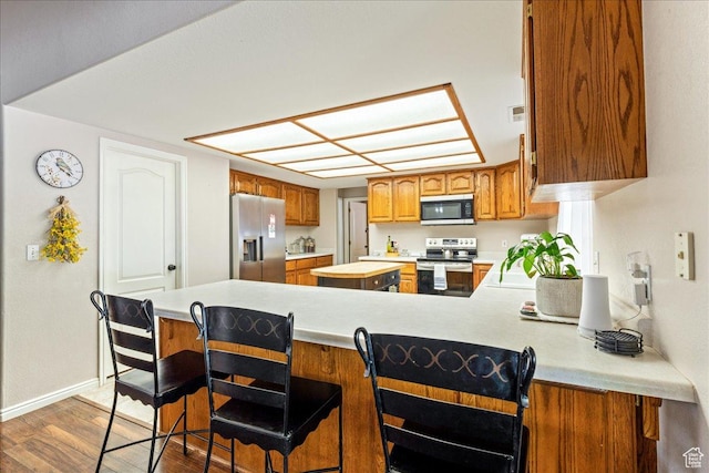 kitchen featuring brown cabinetry, a peninsula, wood finished floors, light countertops, and stainless steel appliances