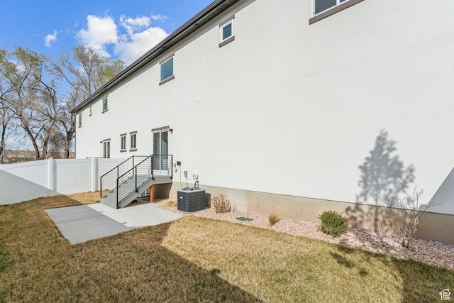 rear view of house with central AC unit, stucco siding, fence, and a lawn