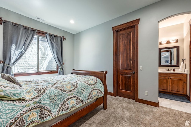 bedroom featuring visible vents, baseboards, arched walkways, ensuite bath, and light colored carpet