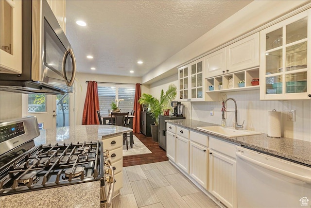 kitchen featuring glass insert cabinets, a sink, appliances with stainless steel finishes, a textured ceiling, and light stone counters