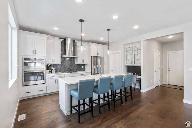 kitchen with wall chimney range hood, light countertops, a kitchen island with sink, a kitchen bar, and stainless steel appliances