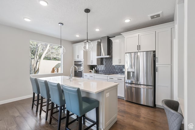 kitchen featuring wall chimney range hood, visible vents, appliances with stainless steel finishes, light countertops, and a center island with sink