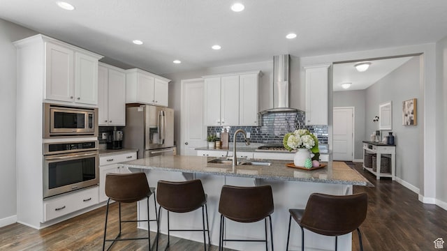 kitchen featuring a kitchen bar, wall chimney range hood, appliances with stainless steel finishes, a sink, and a center island with sink