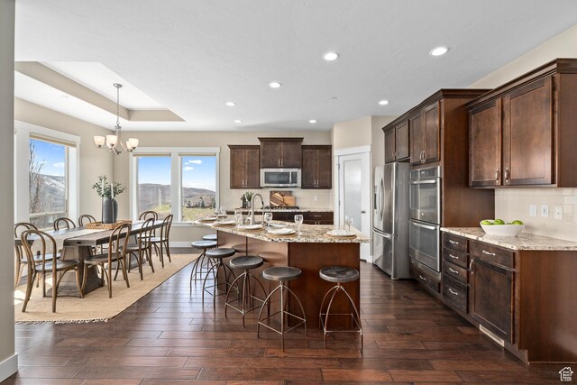 kitchen with dark brown cabinetry, dark wood-style flooring, backsplash, a kitchen island with sink, and appliances with stainless steel finishes