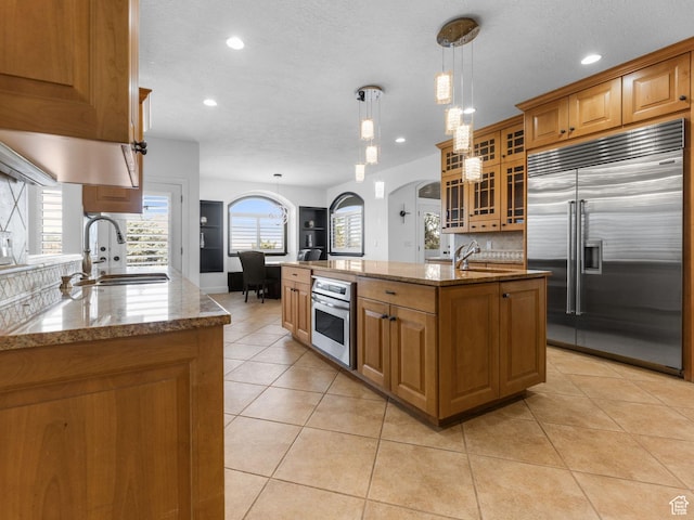 kitchen featuring a sink, brown cabinets, a center island with sink, stone countertops, and stainless steel appliances