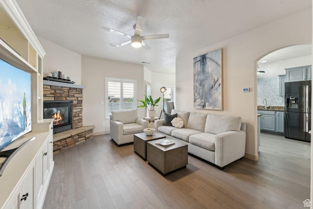 living area featuring ceiling fan, arched walkways, a textured ceiling, a stone fireplace, and light wood finished floors