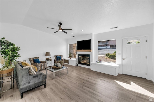 living area with ceiling fan, a glass covered fireplace, vaulted ceiling, visible vents, and wood finished floors