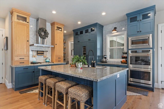 kitchen featuring wall chimney range hood, blue cabinets, a sink, stainless steel appliances, and light wood finished floors