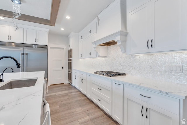 kitchen with built in appliances, light wood-type flooring, premium range hood, backsplash, and white cabinets