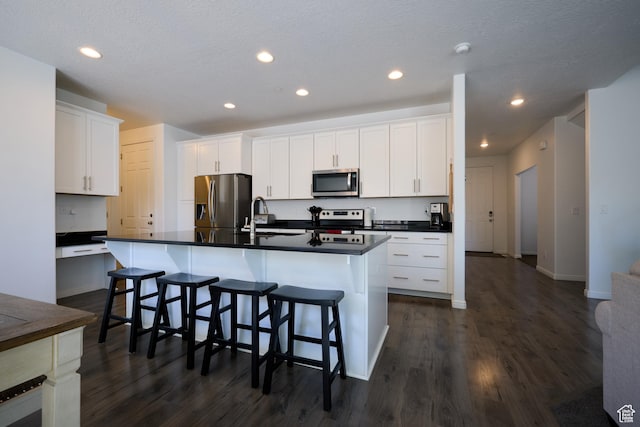 kitchen featuring appliances with stainless steel finishes, dark wood-style flooring, dark countertops, and an island with sink