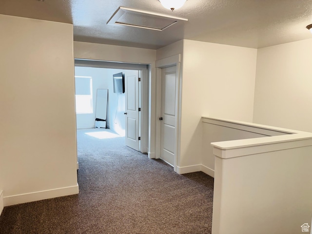 corridor featuring a textured ceiling, attic access, an upstairs landing, dark colored carpet, and baseboards