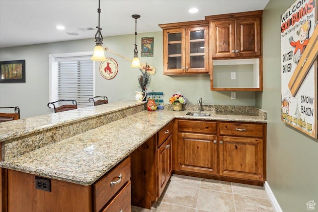kitchen featuring light stone counters, a sink, hanging light fixtures, glass insert cabinets, and a peninsula