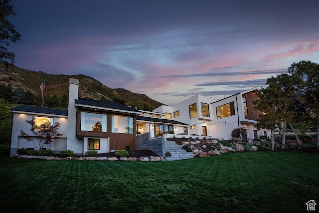 back of house at dusk featuring a chimney, a mountain view, and a lawn