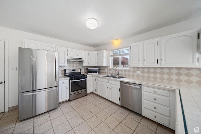 kitchen with white cabinets, a sink, under cabinet range hood, stainless steel appliances, and tile countertops
