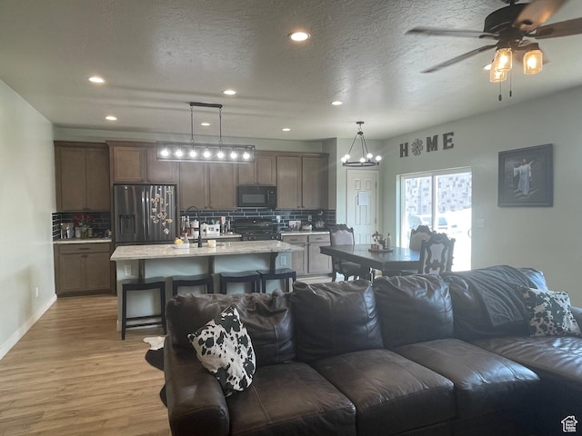 living room featuring ceiling fan with notable chandelier, light wood-style flooring, a textured ceiling, recessed lighting, and baseboards