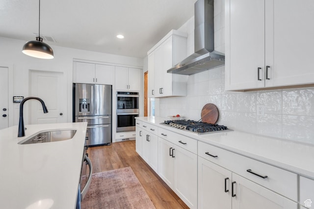 kitchen with wall chimney exhaust hood, light wood-style flooring, appliances with stainless steel finishes, a sink, and backsplash