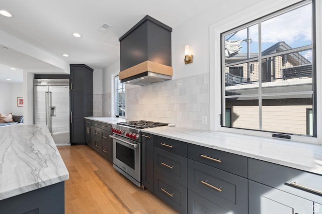 kitchen with light wood-style floors, light stone countertops, premium range hood, premium appliances, and decorative backsplash