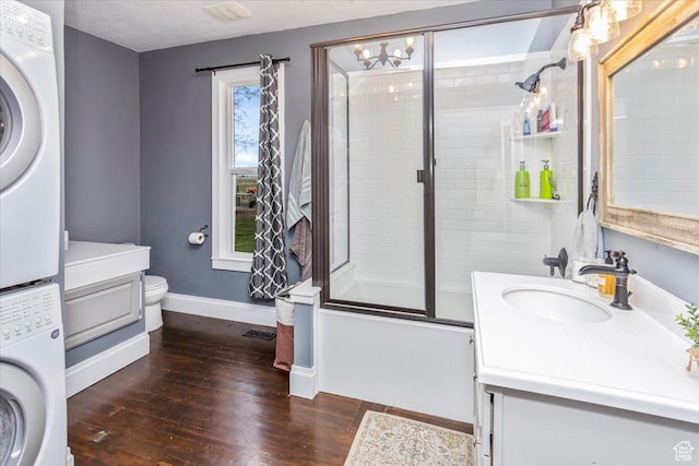 full bath featuring toilet, wood finished floors, enclosed tub / shower combo, stacked washer and clothes dryer, and baseboards