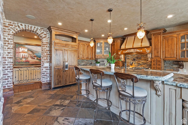13879 S Standing Oak Dr E, Draper, UT 84020 - kitchen with brown cabinetry, stone tile floors, custom range hood, and glass insert cabinets