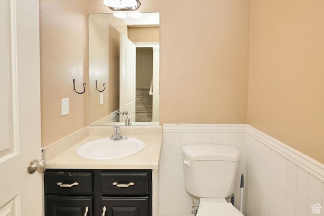 half bath with toilet, a decorative wall, vanity, and wainscoting