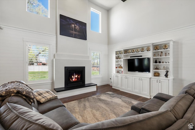 living room with dark wood-style flooring, plenty of natural light, a towering ceiling, and a warm lit fireplace