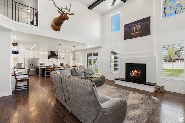 living room featuring dark wood-type flooring, a healthy amount of sunlight, a towering ceiling, and a lit fireplace