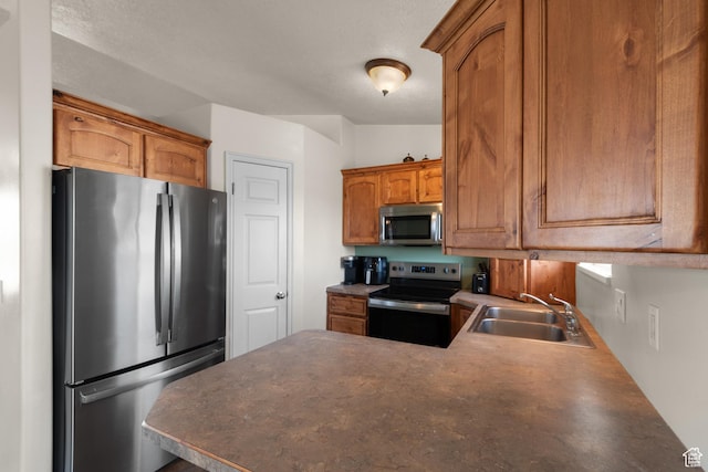 kitchen featuring brown cabinetry, a peninsula, appliances with stainless steel finishes, a sink, and dark countertops