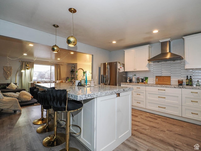 kitchen featuring white cabinetry, stainless steel refrigerator with ice dispenser, light wood-style floors, wall chimney exhaust hood, and a breakfast bar area