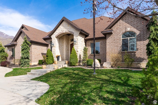 french country inspired facade with brick siding, stone siding, a shingled roof, and a front lawn