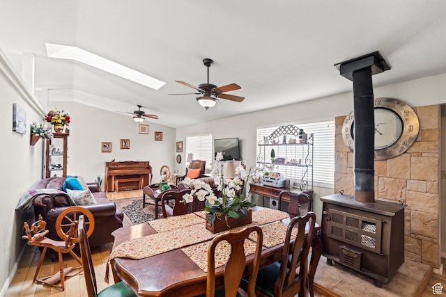 dining area with light wood finished floors, a wood stove, a ceiling fan, and lofted ceiling with skylight