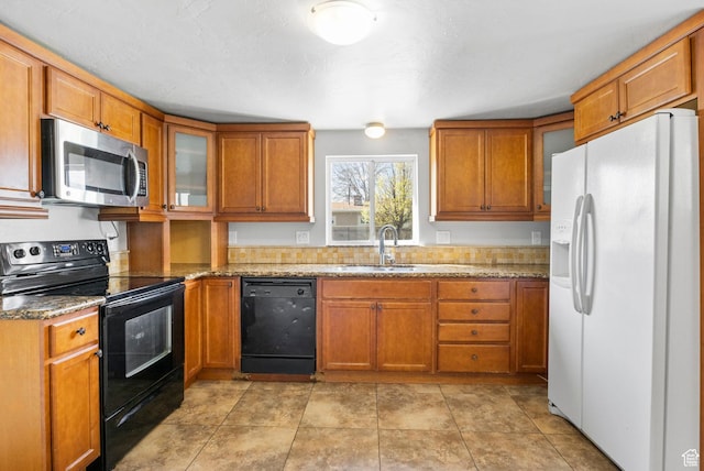 kitchen with light stone counters, glass insert cabinets, black appliances, brown cabinetry, and a sink