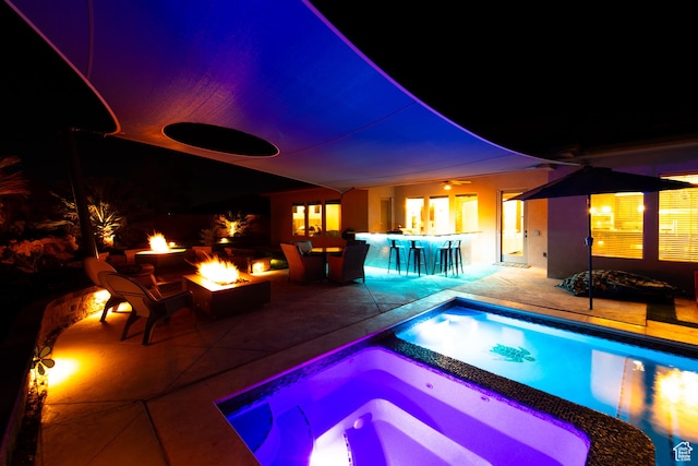 pool at night with a patio, outdoor dry bar, a pool with connected hot tub, and a fire pit